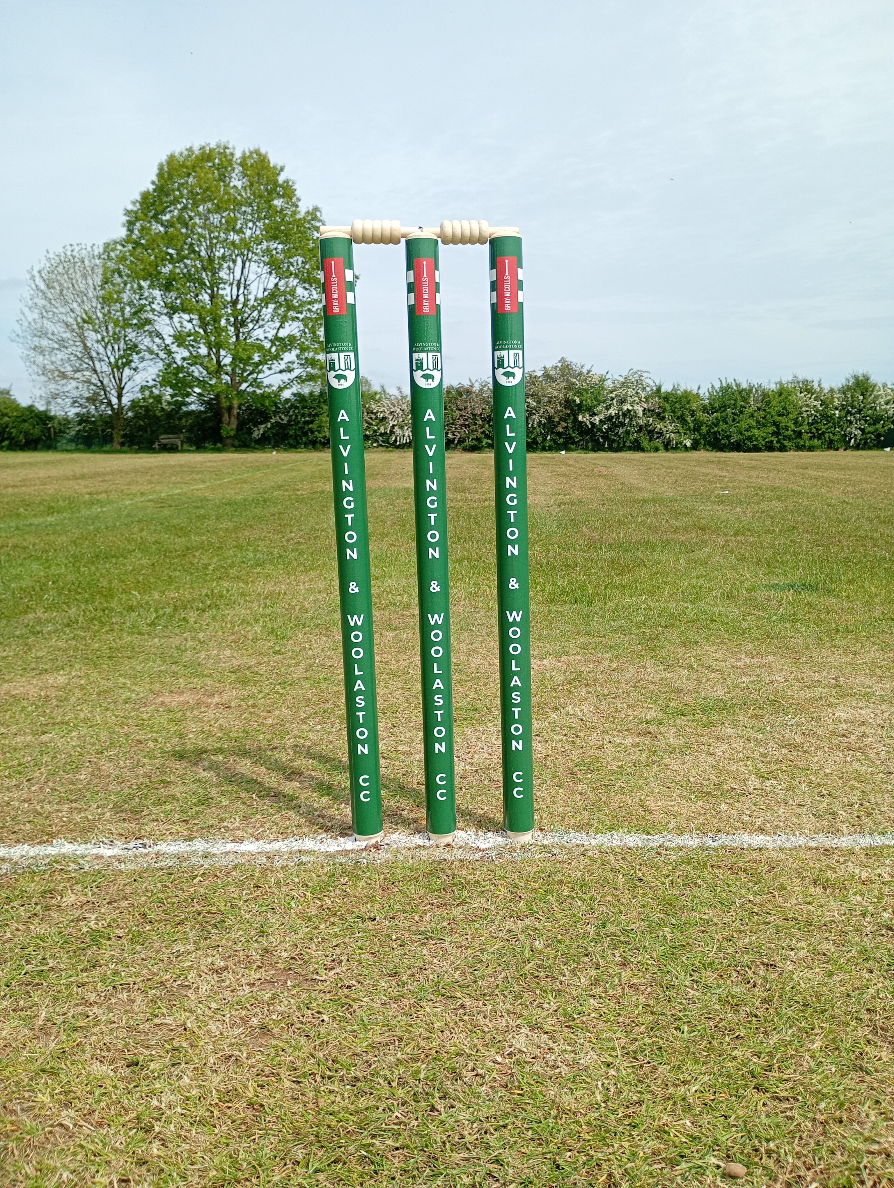 Gray Nicolls Custom Printed Club Cricket Stumps with bails stand on a grassy pitch, featuring a white crease line in front and trees in the background under a clear sky.