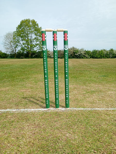 Gray Nicolls Custom Printed Club Cricket Stumps with bails stand on a grassy pitch, featuring a white crease line in front and trees in the background under a clear sky.
