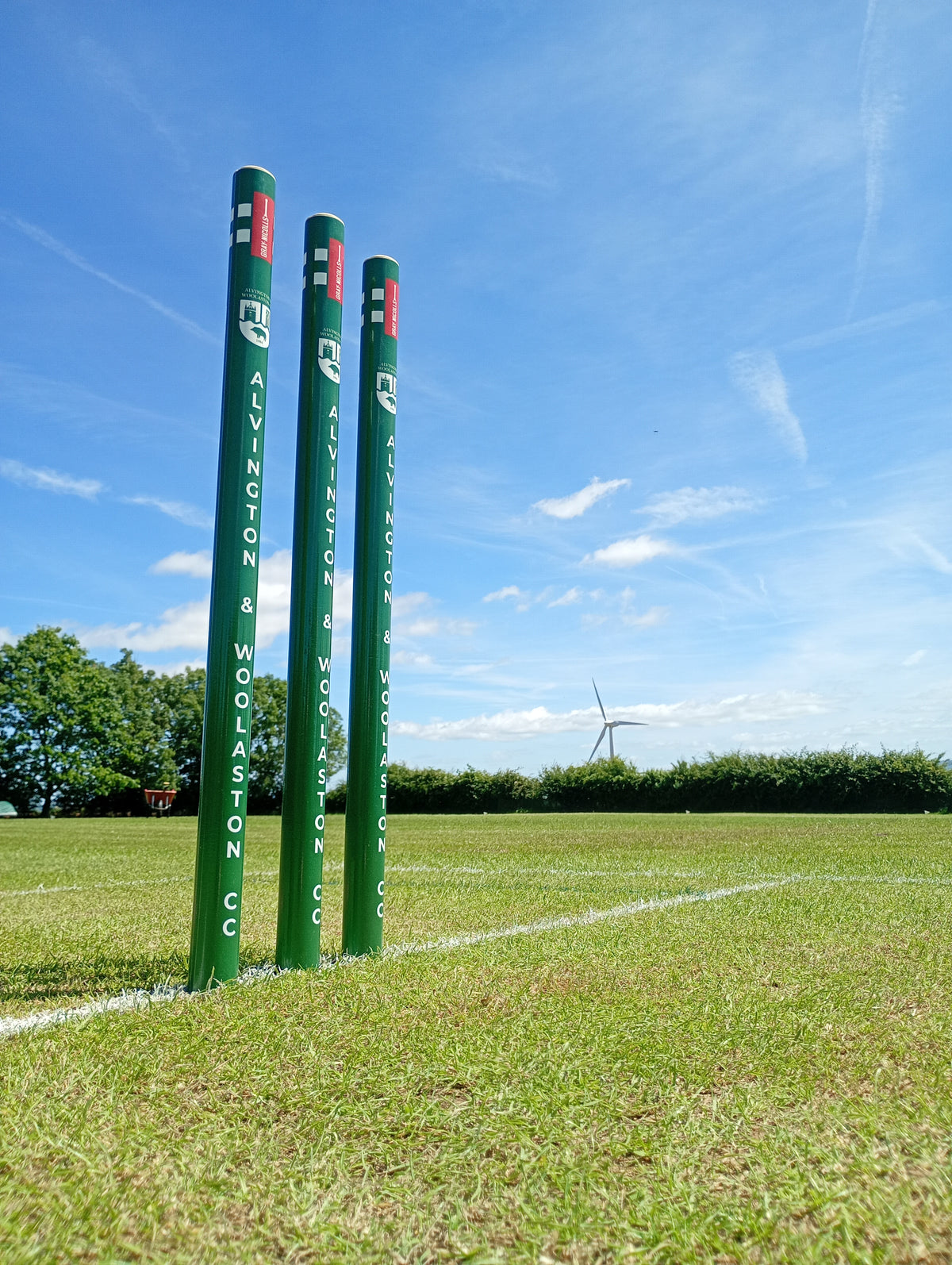 Three green Gray Nicolls Custom Printed Club Cricket Stumps stand on a grassy field under a blue sky, with a wind turbine and trees visible in the background.