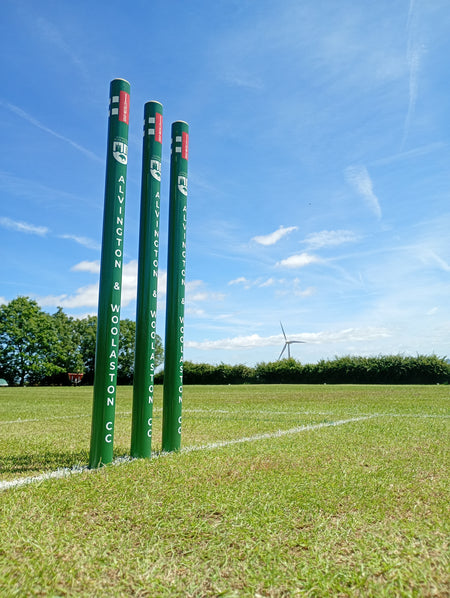 Three green Gray Nicolls Custom Printed Club Cricket Stumps stand on a grassy field under a blue sky, with a wind turbine and trees visible in the background.