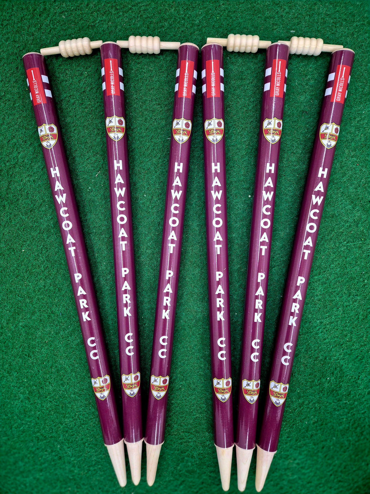 Six maroon Gray Nicolls Custom Printed Club Cricket Stumps, each featuring your club name and crest, arranged on green turf with bails on top—these stumps add a professional touch to any cricket setup.