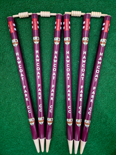 Six maroon Gray Nicolls Custom Printed Club Cricket Stumps, each featuring your club name and crest, arranged on green turf with bails on top—these stumps add a professional touch to any cricket setup.