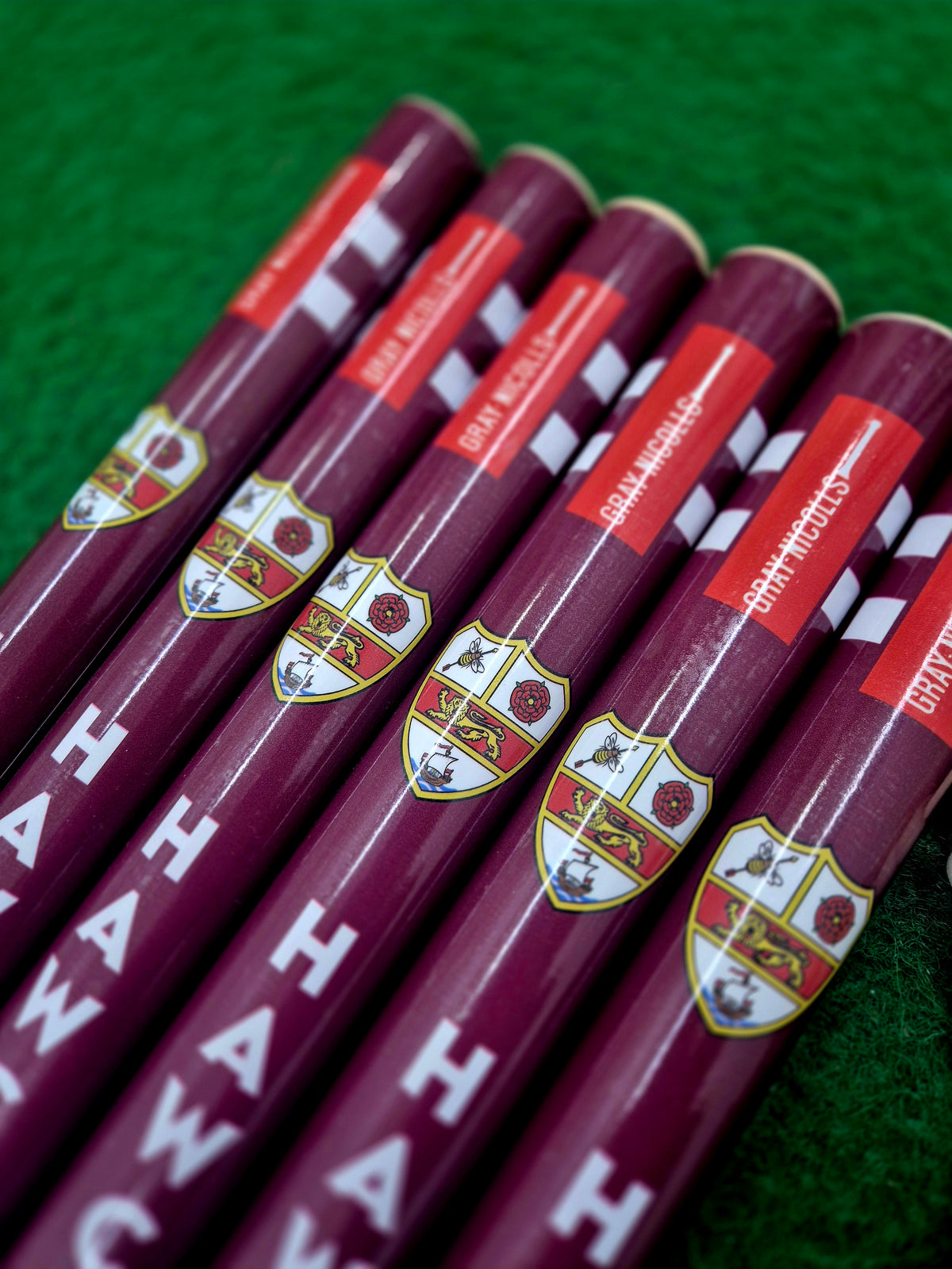 Six maroon cricket bats with crest logos and red labels are laid out on green grass beside Gray Nicolls Custom Printed Club Cricket Stumps, giving the scene a club equipment vibe with branded text and symbols visible.
