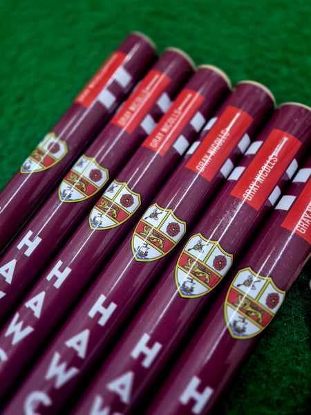 Six maroon cricket bats with crest logos and red labels are laid out on green grass beside Gray Nicolls Custom Printed Club Cricket Stumps, giving the scene a club equipment vibe with branded text and symbols visible.