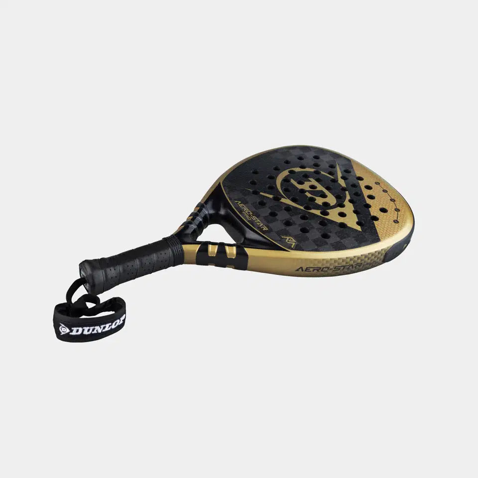 The Dunlop Aerostar Pro Padel Racket by Dunlop features a black and gold design, Power Holes, a perforated surface, 16K Carbon construction, textured grip, and an attached wrist strap—shown on a plain light background.