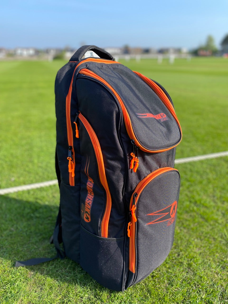 A black and orange Katchet KoachSak One2One bag with multiple zippered compartments stands on a grassy sports field, with blurred white goalposts and buildings in the background.