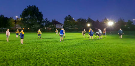 A group plays rugby on a grassy field at dusk under Ritelite Sports Lite Portable Floodlights, with trees and houses in the background. Some players wear blue and yellow jerseys.