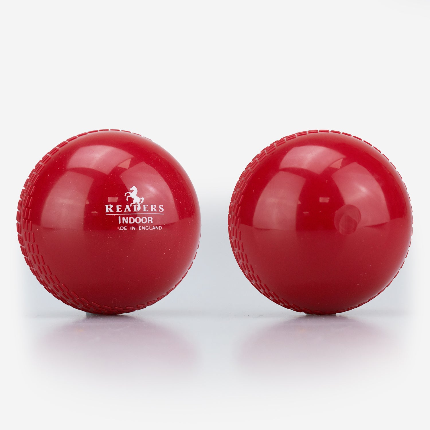 Two shiny red Readers Indoor 4oz Cricket Balls lie side by side on a white surface—one features the Readers logo, while the other highlights its smooth, lightweight plastic design, perfect for indoor training or coaching.