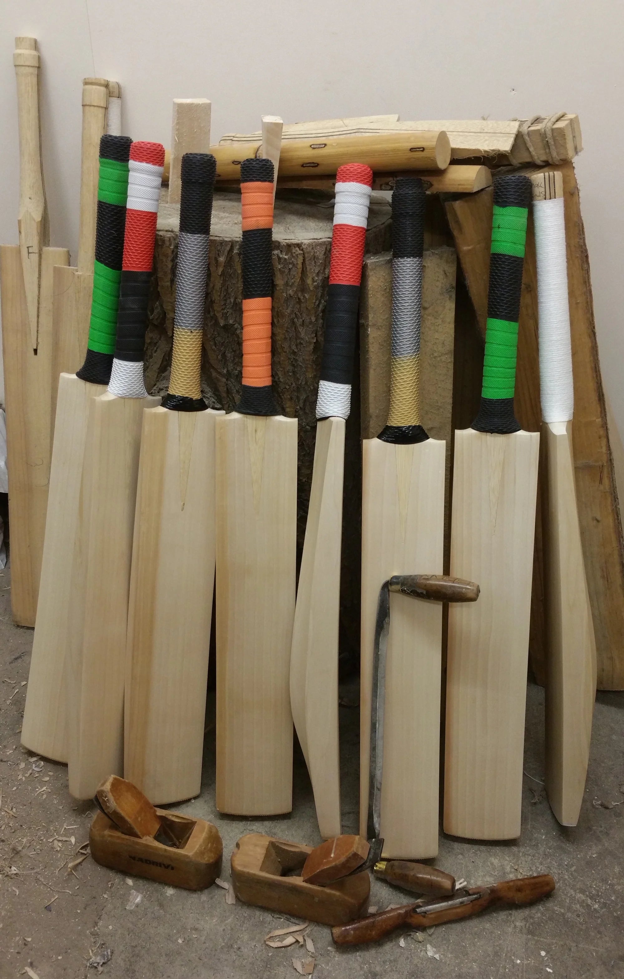 Six cricket bats with colorful grips, ready for the Cricket Bat Repair - Full Refurbishment by Martin Berrill Sports, lean against a log amid unfinished bats, tools, and wood shavings.