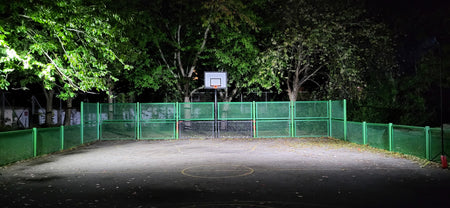 At night, a small outdoor basketball court with one hoop, green fence, and trees is brightly lit by the Nightsearcher SportStar Portable LED Floodlight (20,000 lumens), creating a vibrant nighttime scene.