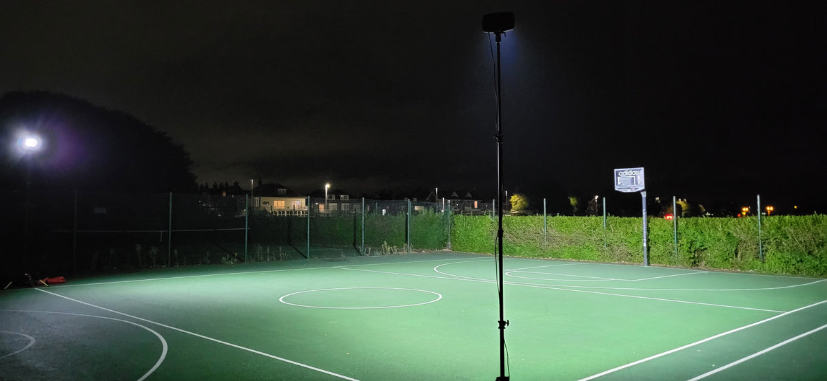 A well-lit outdoor basketball court at night, illuminated by Nightsearcher SportStar Portable LED Floodlights, features a hoop and hedge in the background with houses and streetlights visible beyond the court.