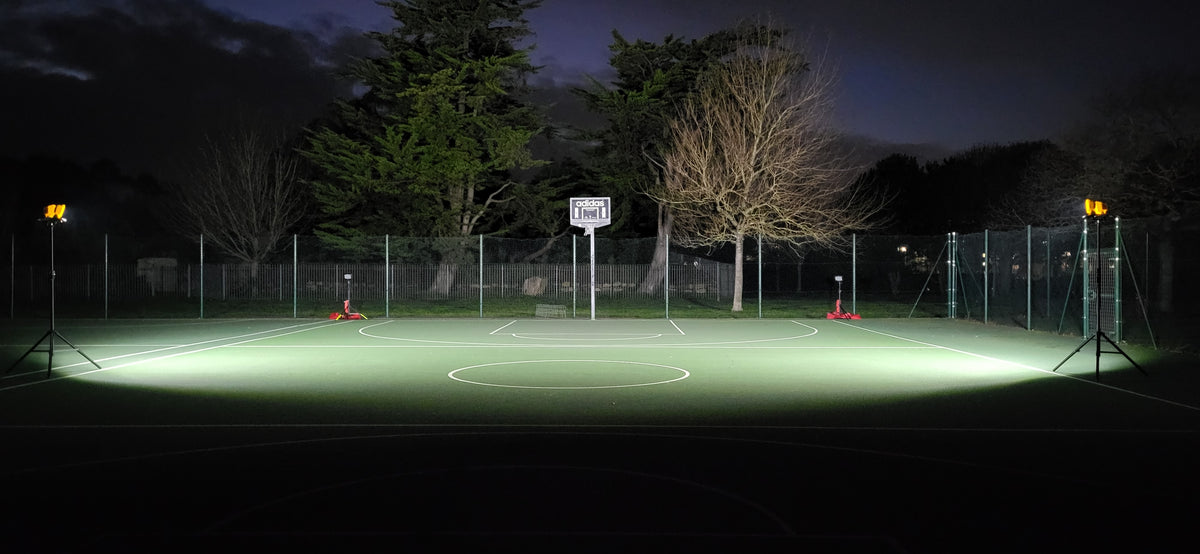 At night, a basketball court is lit by four Nightsearcher Galaxy Pro Twin Portable Floodlights at the corners. Trees and a wire fence are visible in the darkened background.