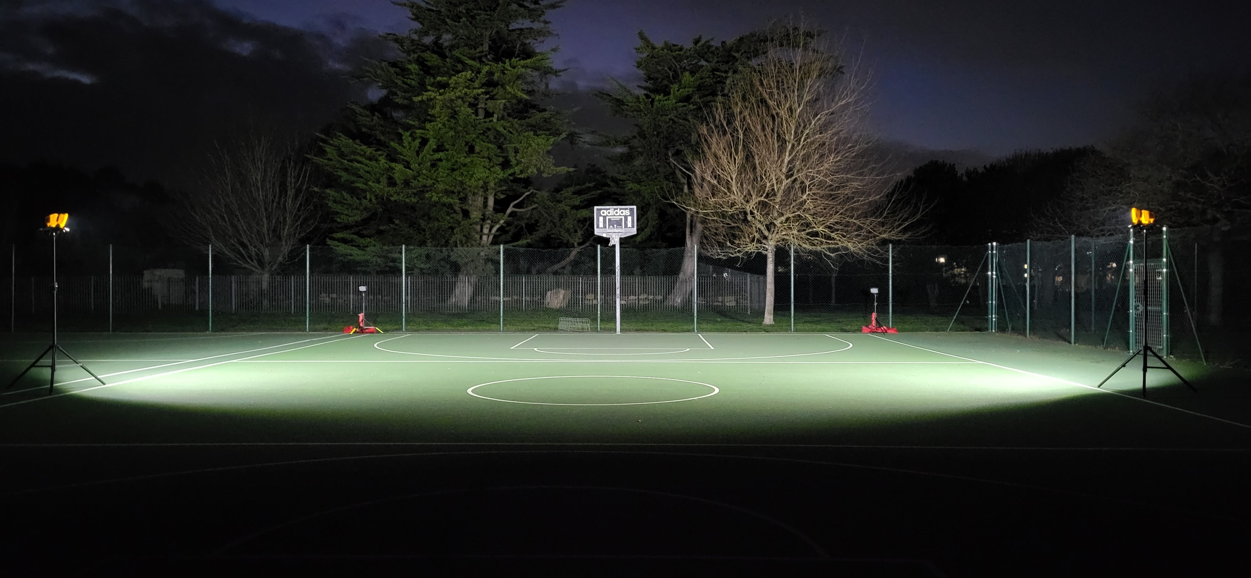 At night, a basketball court is lit by four Nightsearcher Galaxy Pro Twin Portable Floodlights at the corners. Trees and a wire fence are visible in the darkened background.