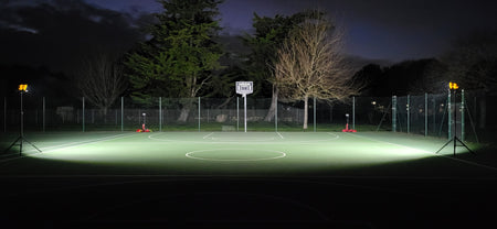 At night, a basketball court is lit by four Nightsearcher Galaxy Pro Twin Portable Floodlights at the corners. Trees and a wire fence are visible in the darkened background.
