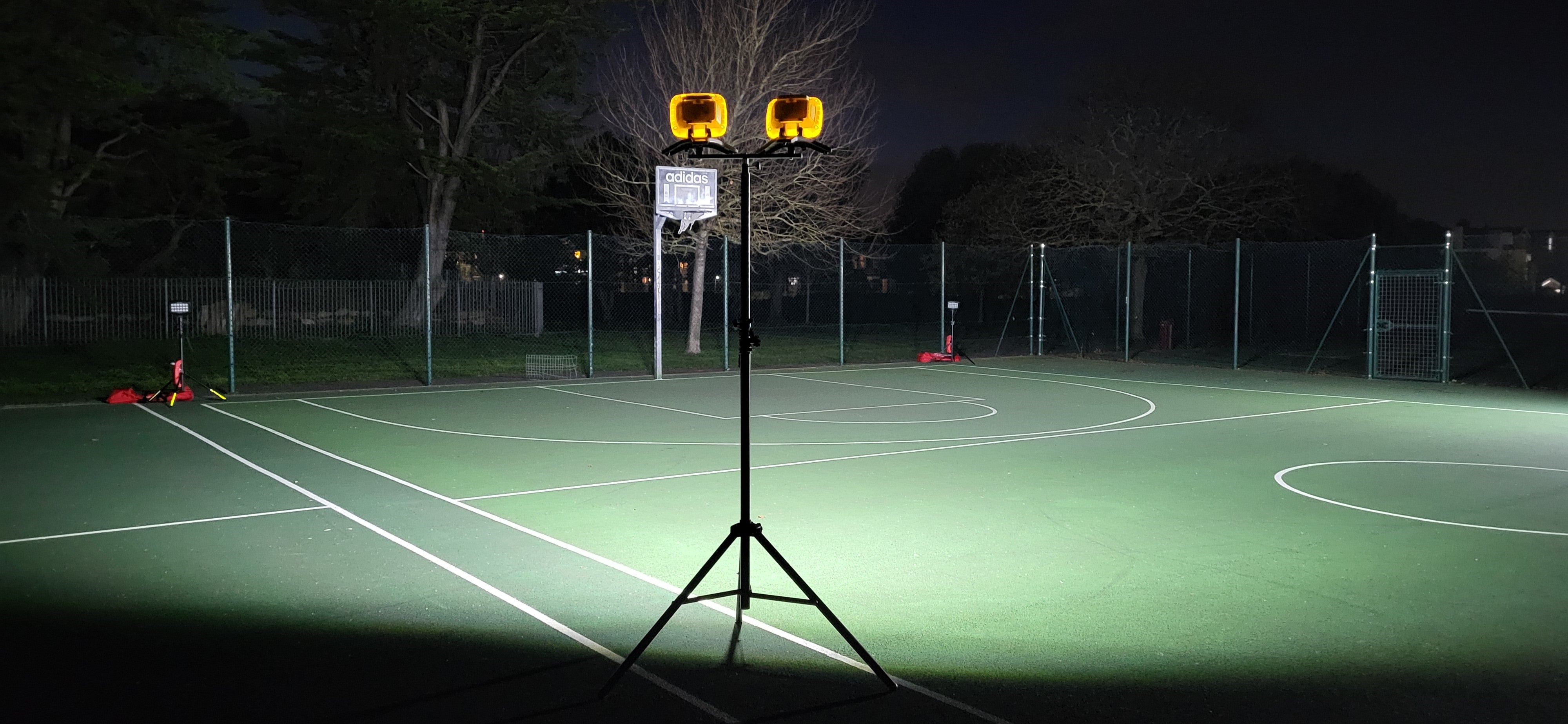 The Nightsearcher Galaxy Pro Twin Portable Floodlights on a tripod brightly illuminate an empty outdoor tennis court at night, surrounded by a wire fence and trees.