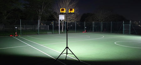 The Nightsearcher Galaxy Pro Twin Portable Floodlights on a tripod brightly illuminate an empty outdoor tennis court at night, surrounded by a wire fence and trees.