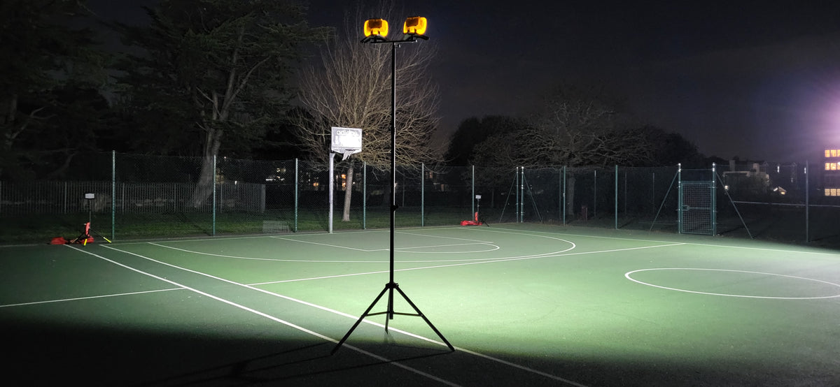 A sports court at night is brightly lit by the Nightsearcher Galaxy Pro Twin Portable Floodlights on a tripod. With trees and a fence in the dark background, it’s ideal for outdoor training light setups.
