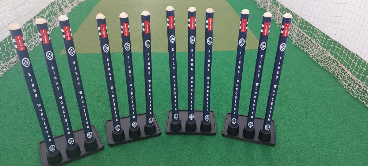 Several sets of Gray Nicolls Custom Printed Match Cricket Stumps with Heavy Rubber Base, featuring a white logo and KINGS WORCESTER branding, stand upright on green artificial turf inside an indoor cricket net area.