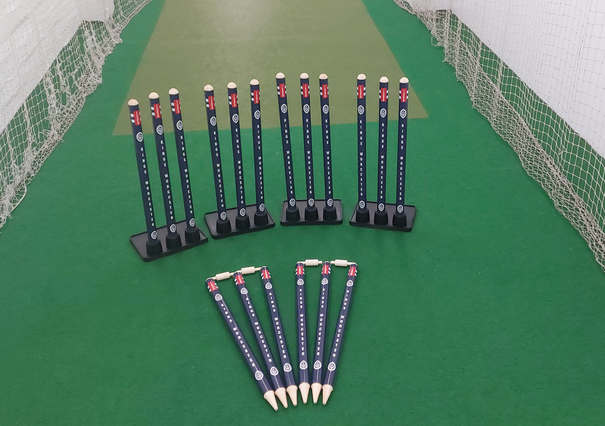 Three Gray Nicolls Custom Printed Match Cricket Stumps with Heavy Rubber Base and six cricket-themed pencils are neatly arranged on green artificial turf in an indoor practice net area.