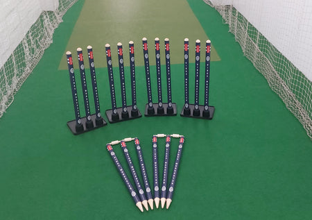 Three Gray Nicolls Custom Printed Match Cricket Stumps with Heavy Rubber Base and six cricket-themed pencils are neatly arranged on green artificial turf in an indoor practice net area.