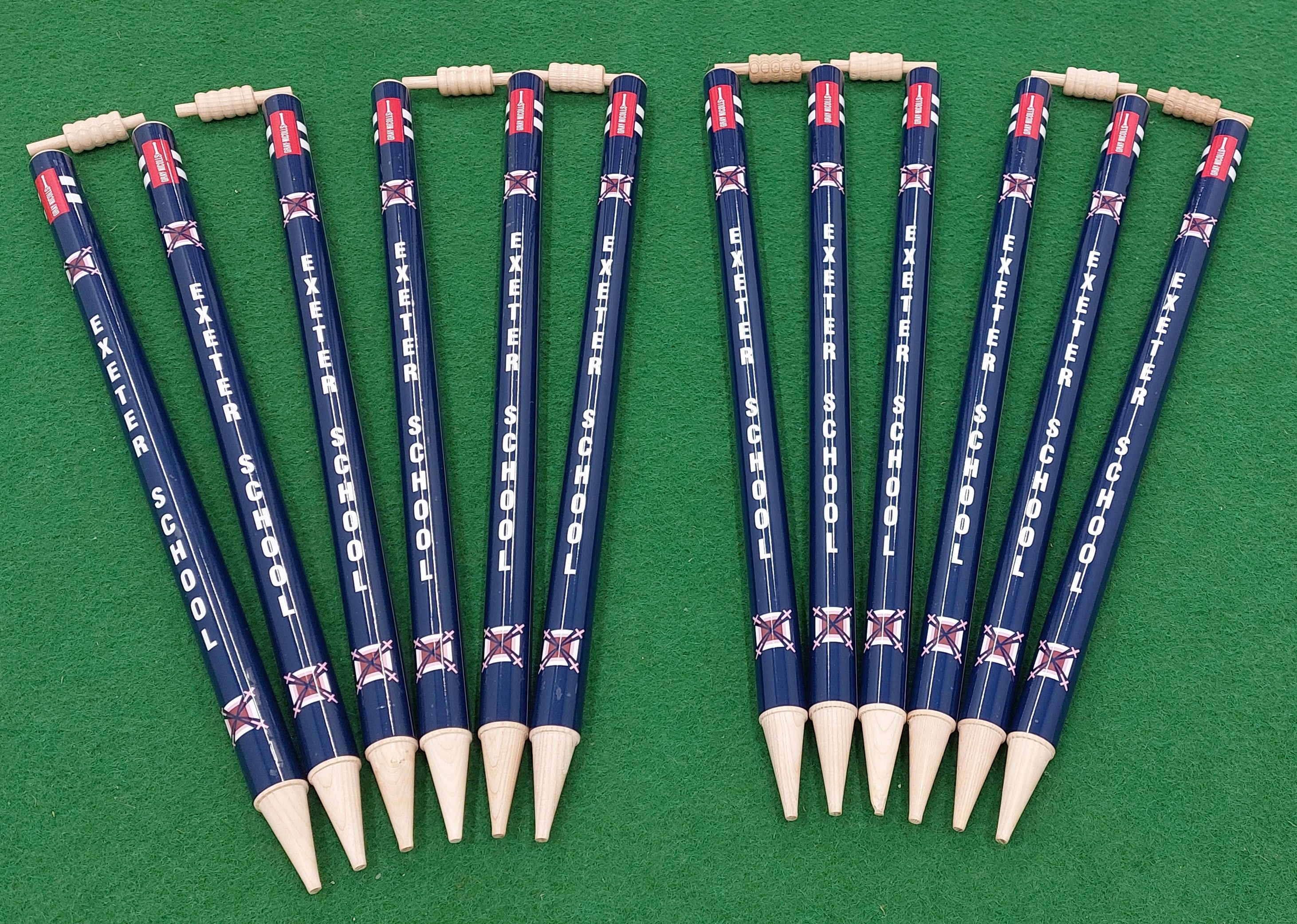 Ten Gray Nicolls Custom Printed Club Cricket Stumps, arranged in two neat groups on a green field, stand out with their bold design and personalized branding. Each stump features classic detailing and represents Exeter School.