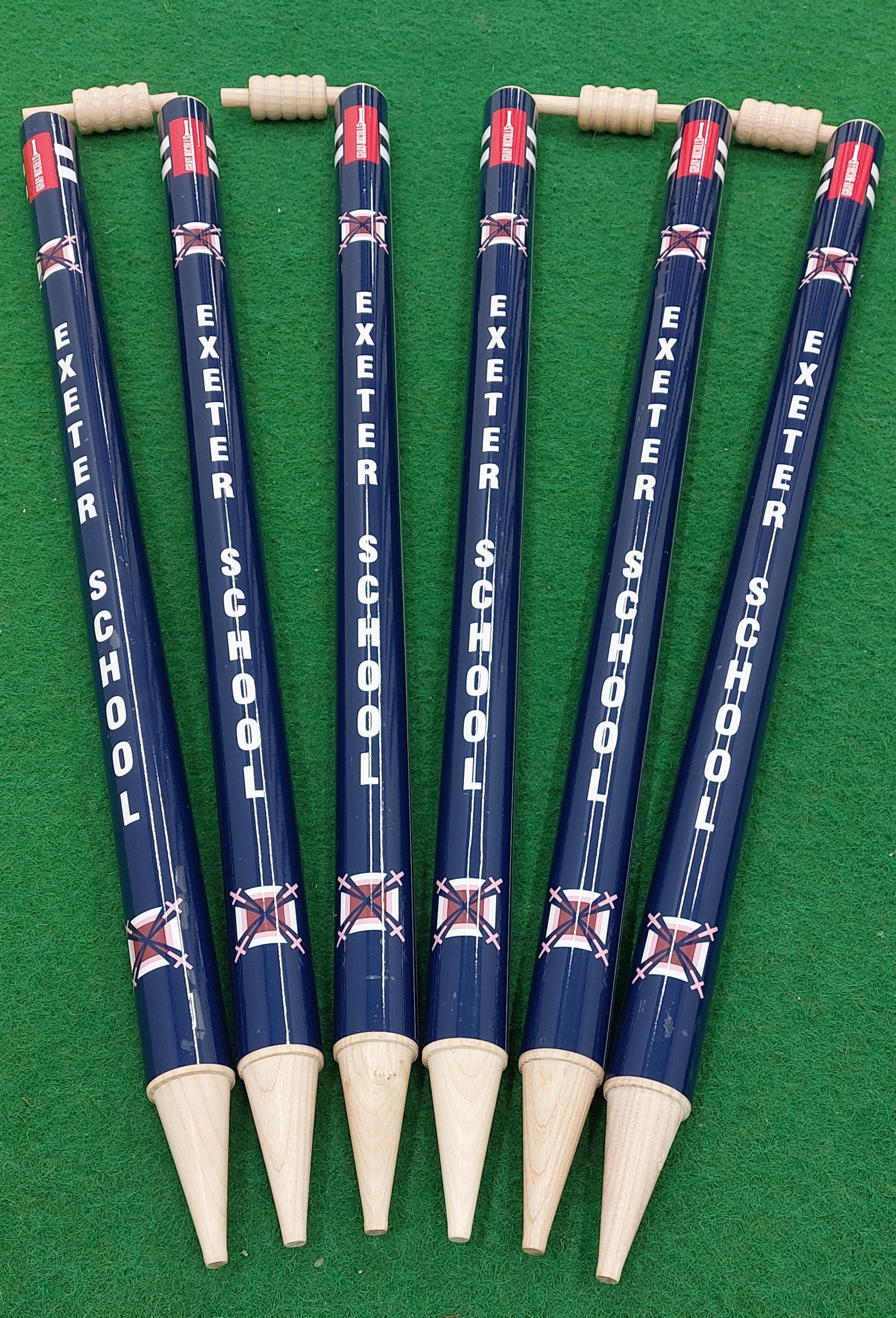 Six blue and wooden Gray Nicolls Custom Printed Club Cricket Stumps, featuring EXETER SCHOOL printed vertically in white, each topped with a wooden peg and arranged side by side on green artificial grass.