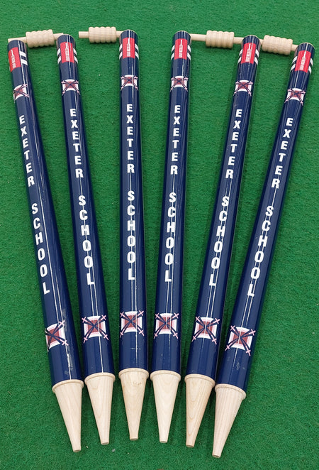 Six blue and wooden Gray Nicolls Custom Printed Club Cricket Stumps, featuring EXETER SCHOOL printed vertically in white, each topped with a wooden peg and arranged side by side on green artificial grass.