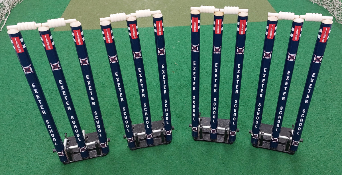 Four sets of Custom Printed Spring Back Stumps by Gray Nicolls with white bails on a green court. Each set has three cricket stumps, sturdy base braces for stability, and is labeled EXETER SCHOOL.