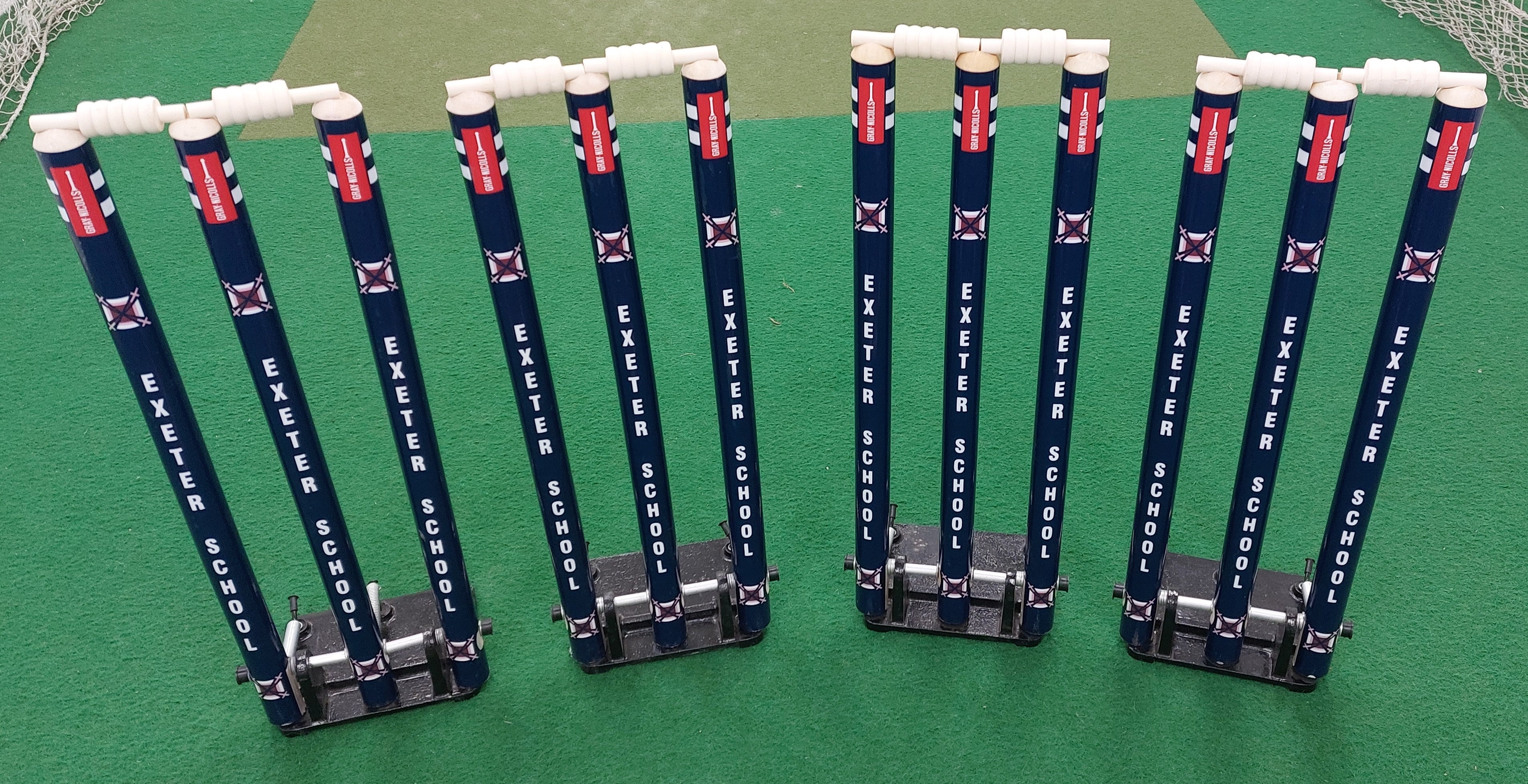 Four sets of Custom Printed Spring Back Stumps by Gray Nicolls with white bails on a green court. Each set has three cricket stumps, sturdy base braces for stability, and is labeled EXETER SCHOOL.