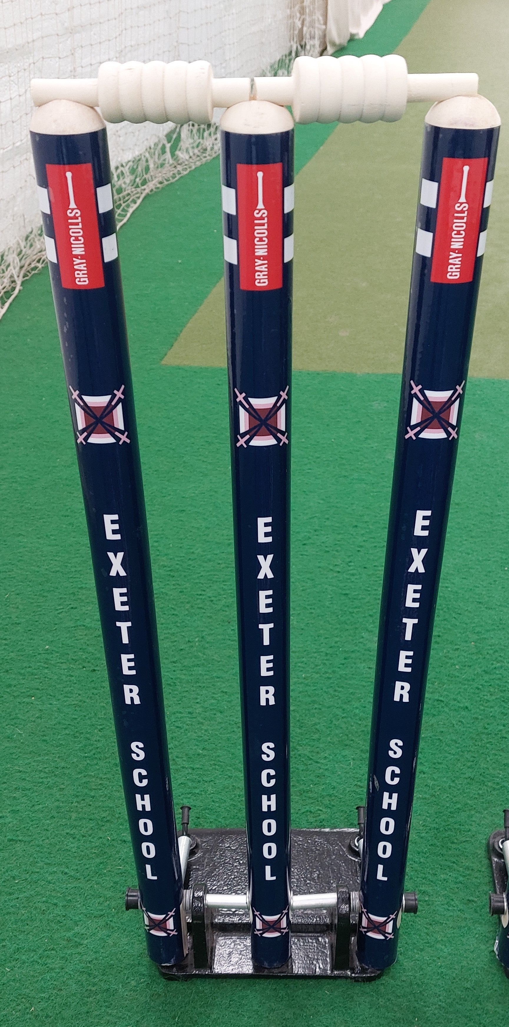 Three blue Gray Nicolls Custom Printed Spring Back Stumps labeled Exeter School, with white bails, are set on a green artificial pitch in an indoor cricket training area—ideal for practice sessions.