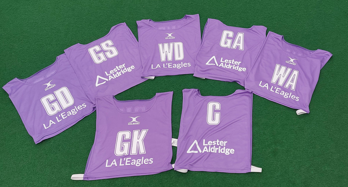 Seven purple Personalised Gilbert Netball Bibs (Reversible) by Gilbert, each with white player positions (GD, GS, WD, GA, WA, GK, C), and LA LEagles and Lester Aldridge logos laid out on a green surface.