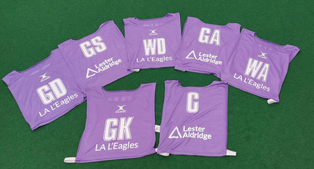 Seven purple Personalised Gilbert Netball Bibs (Reversible) by Gilbert, each with white player positions (GD, GS, WD, GA, WA, GK, C), and LA LEagles and Lester Aldridge logos laid out on a green surface.