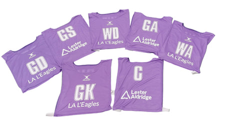 Seven purple Personalised Gilbert Netball Bibs by Gilbert are arranged in a semicircle. Each displays a white position (GS, GA, WA, WD, GD, GK, C), plus LA LEagles and sponsor Lester Aldridge.
