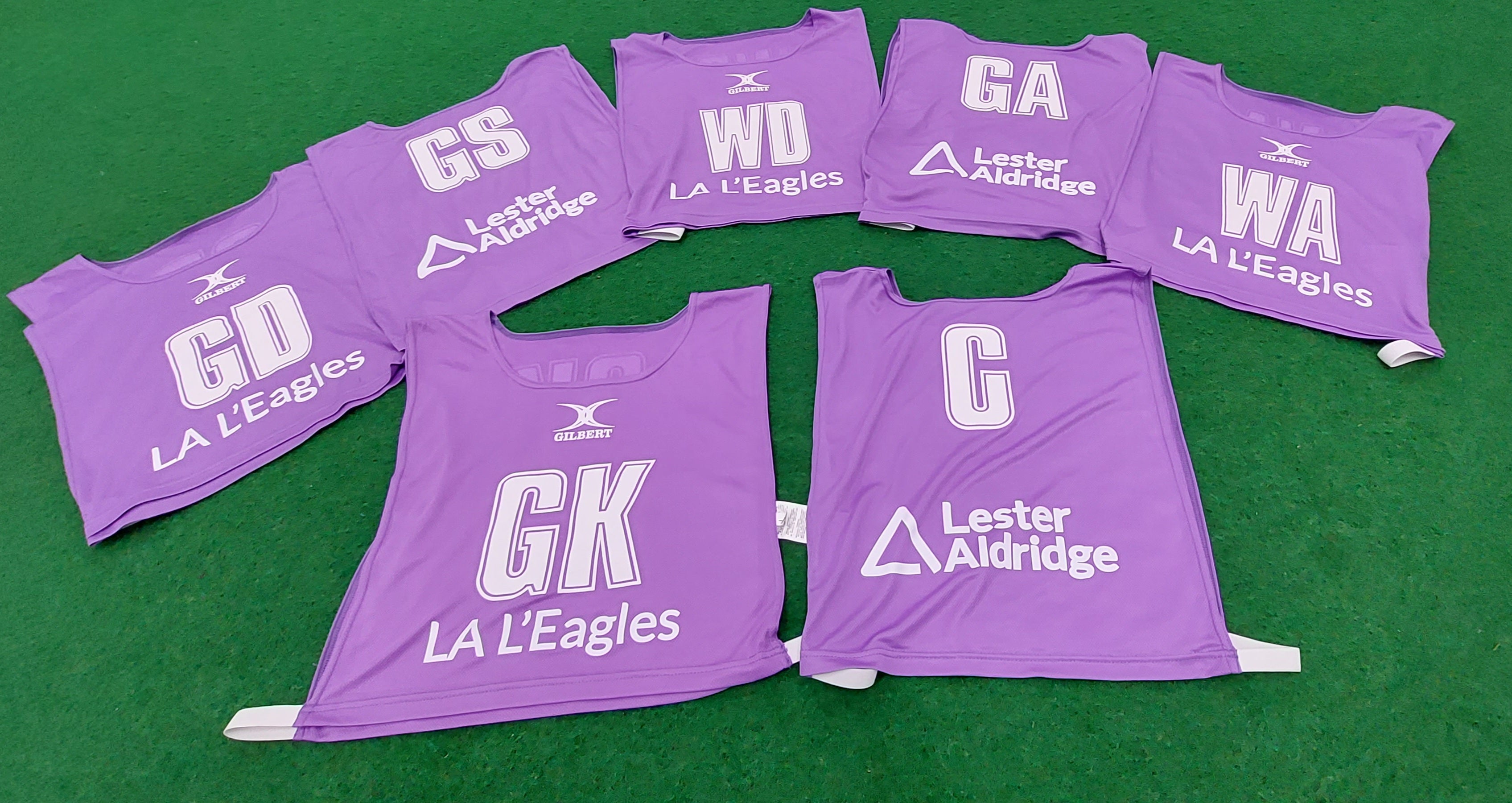 Seven purple Personalised Gilbert Netball Bibs (Reversible) are displayed on green turf, each marked with a unique position (GS, GA, WA, C, WD, GD, GK) and Gilbert branding.