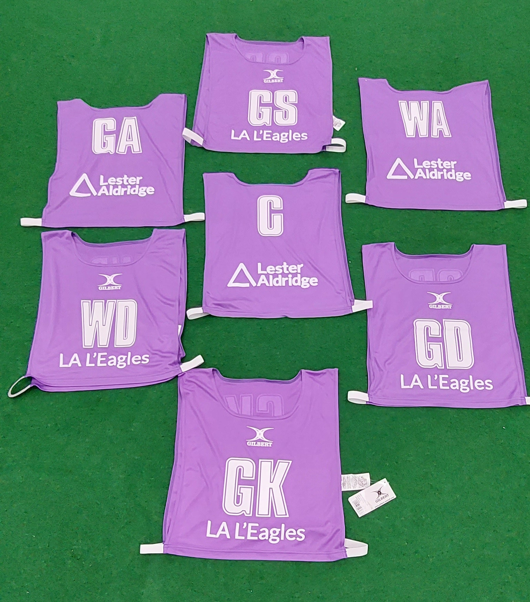 Seven purple Personalised Gilbert Netball Bibs by Gilbert, each showing a netball position (GA, GS, WA, C, WD, GK, GD), plus “LA L’Eagles” and the Lester Aldridge sponsor logo, are arranged on a green floor.