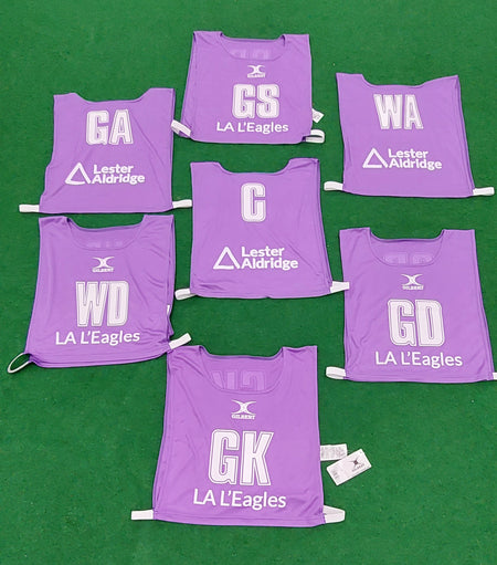 Seven purple Personalised Gilbert Netball Bibs by Gilbert, each showing a netball position (GA, GS, WA, C, WD, GK, GD), plus “LA L’Eagles” and the Lester Aldridge sponsor logo, are arranged on a green floor.