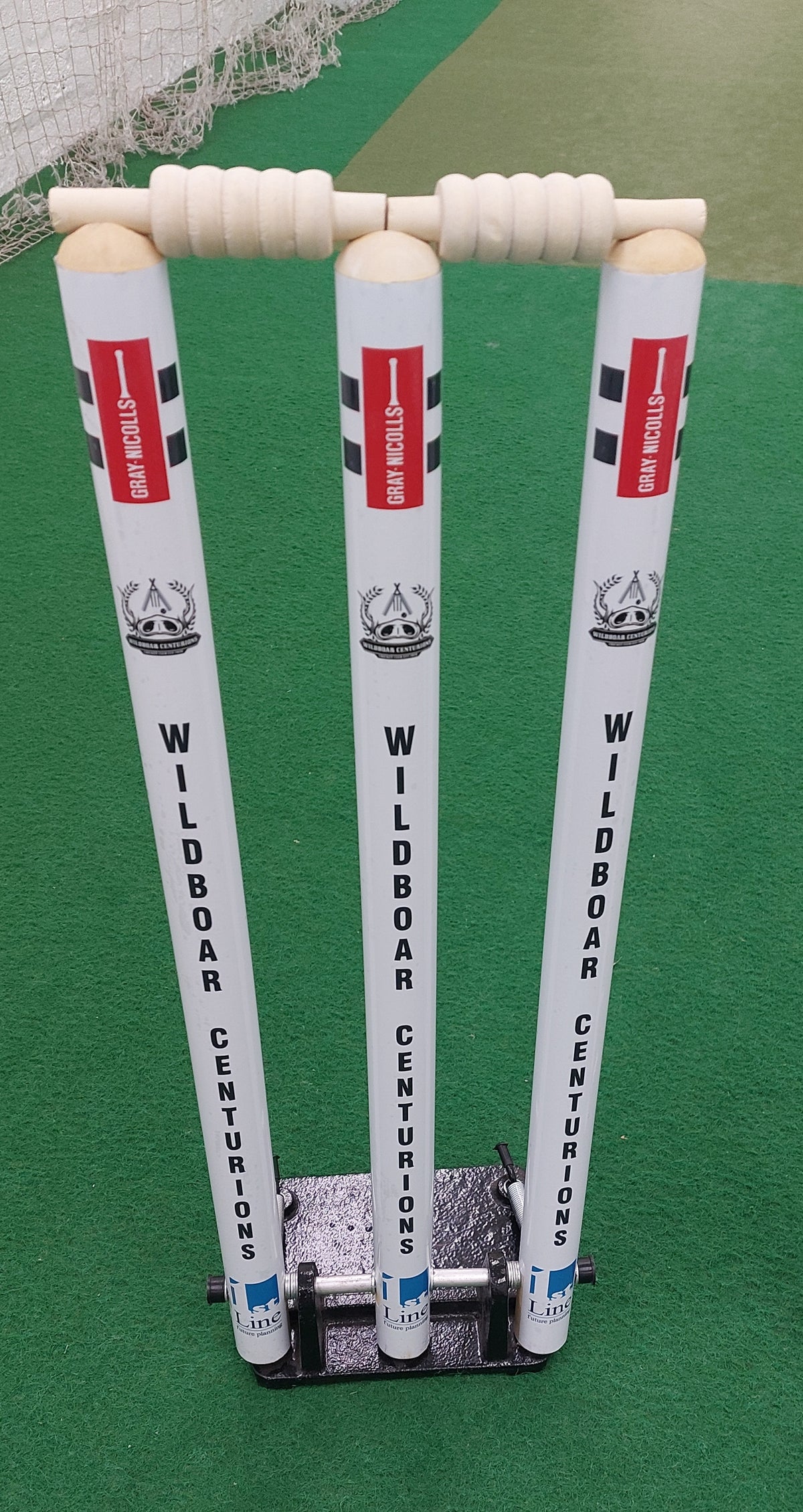 Three white Gray Nicolls Custom Printed Spring Back Stumps, featuring "WILDBOAR CENTURIONS" text and two bails, stand on a green artificial pitch. These spring-loaded stumps are fixed in a sturdy black base for cricket training sessions.