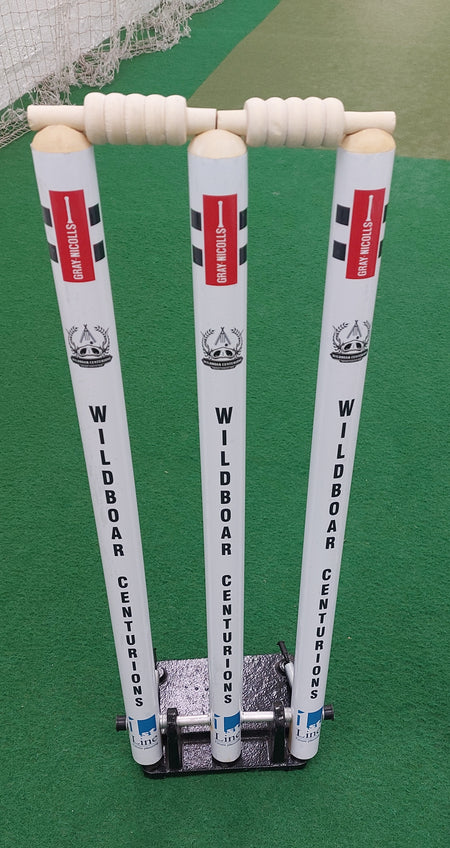 Three white Gray Nicolls Custom Printed Spring Back Stumps, featuring "WILDBOAR CENTURIONS" text and two bails, stand on a green artificial pitch. These spring-loaded stumps are fixed in a sturdy black base for cricket training sessions.