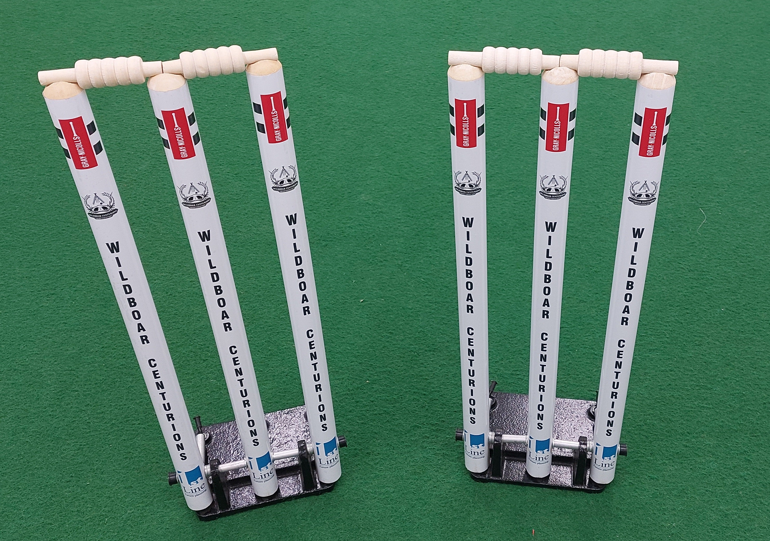 Two sets of Custom Printed Spring Back Stumps by Gray Nicolls, in white with bails, stand upright on black bases atop a green field—perfect as spring-loaded cricket training stumps.