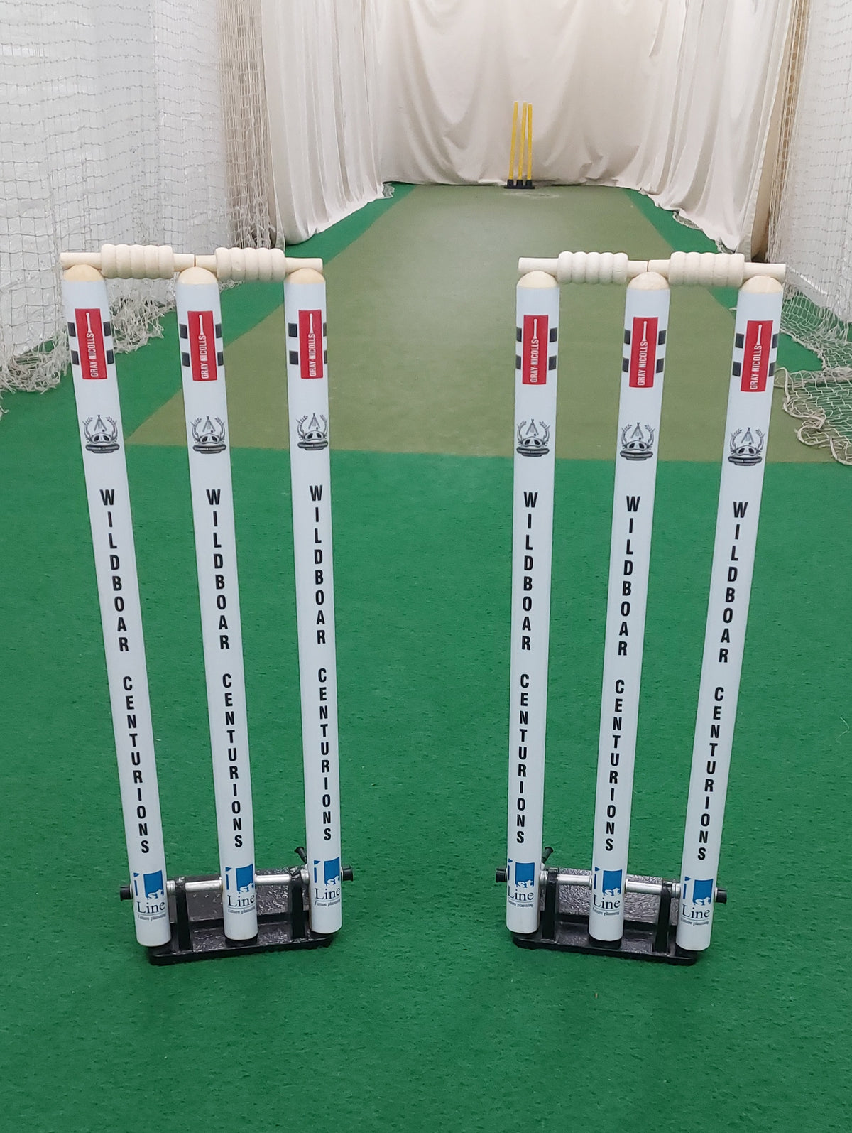 Two sets of Gray Nicolls Custom Printed Spring Back Stumps stand on artificial green turf in an indoor net, ideal for consistent, durable cricket training. Another set is visible in the background.