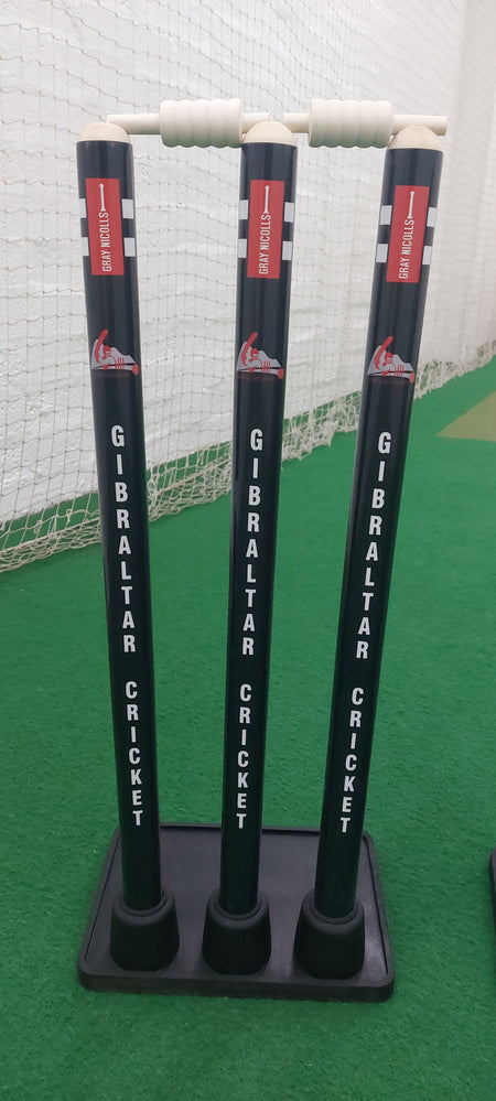 Three Gray Nicolls Custom Printed Match Cricket Stumps with heavy rubber bases stand on a green indoor pitch, each topped with white bails, and a white net in the background.
