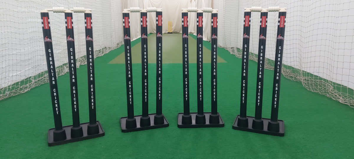 Three sets of Gray Nicolls Custom Printed Match Cricket Stumps with Heavy Rubber Base and white bails stand on green artificial turf inside an indoor practice net.