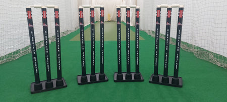 Three sets of Gray Nicolls Custom Printed Match Cricket Stumps with Heavy Rubber Base and white bails stand on green artificial turf inside an indoor practice net.