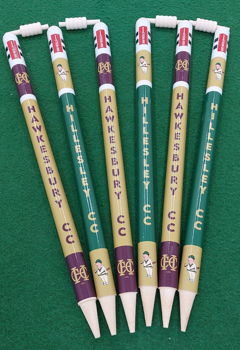Six Gray Nicolls Custom Printed Club Cricket Stumps, in green and gold with cricket player graphics and erasers on top, are arranged fan-shaped on a green surface to resemble souvenir pencils labeled HAWKESBURY CC and HILLESLEY CC.