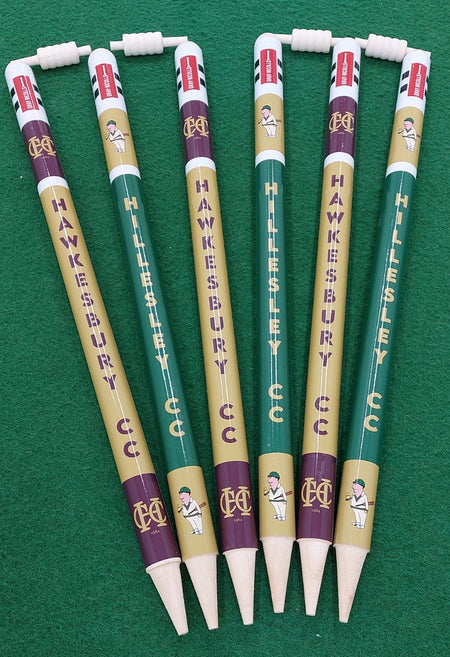 Six Gray Nicolls Custom Printed Club Cricket Stumps, in green and gold with cricket player graphics and erasers on top, are arranged fan-shaped on a green surface to resemble souvenir pencils labeled HAWKESBURY CC and HILLESLEY CC.