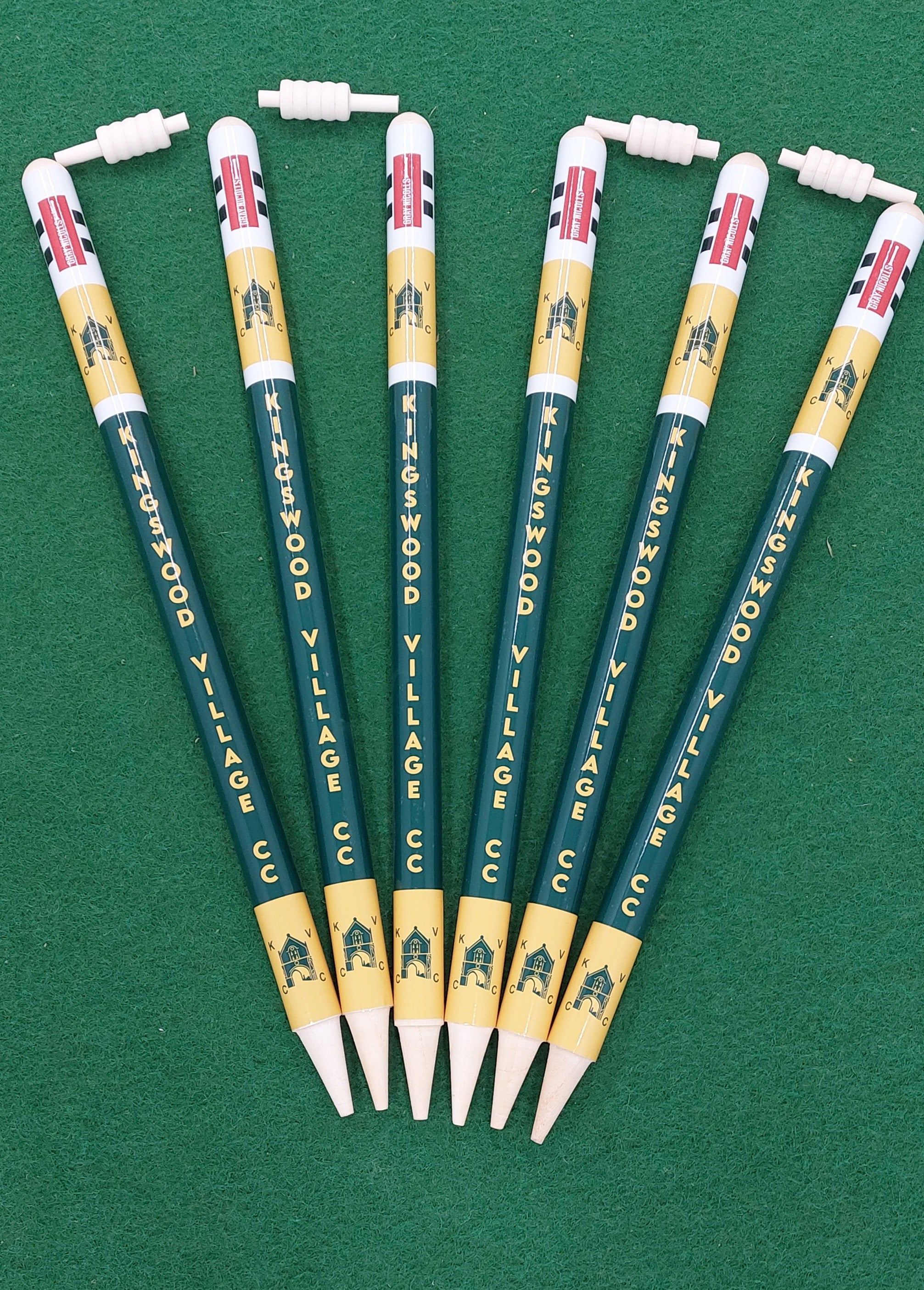 Six Gray Nicolls Custom Printed Club Cricket Stumps pens, with KINGSWOOD VILLAGE CC printed on them, are arranged fan-shaped on a green surface. The yellow and green pens have clip-on toppers shaped like cricket stumps, bats, and wickets.