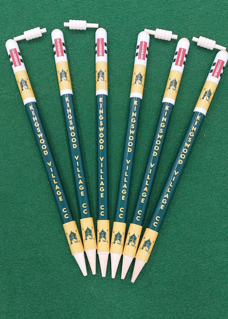 Six Gray Nicolls Custom Printed Club Cricket Stumps pens, with KINGSWOOD VILLAGE CC printed on them, are arranged fan-shaped on a green surface. The yellow and green pens have clip-on toppers shaped like cricket stumps, bats, and wickets.