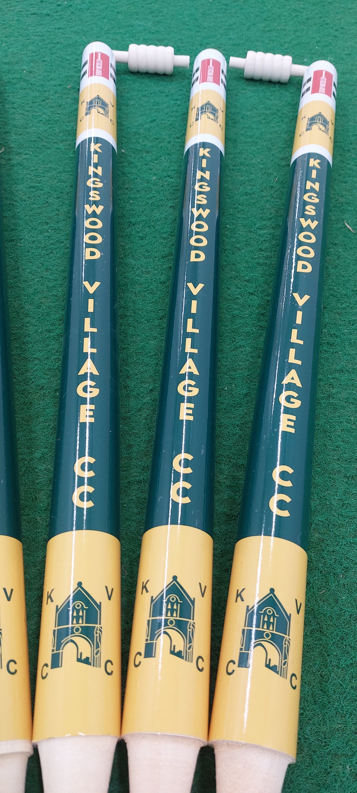Four Gray Nicolls Custom Printed Club Cricket Stumps featuring the Kingswood Village CCC name and building logo, displayed on a green surface.