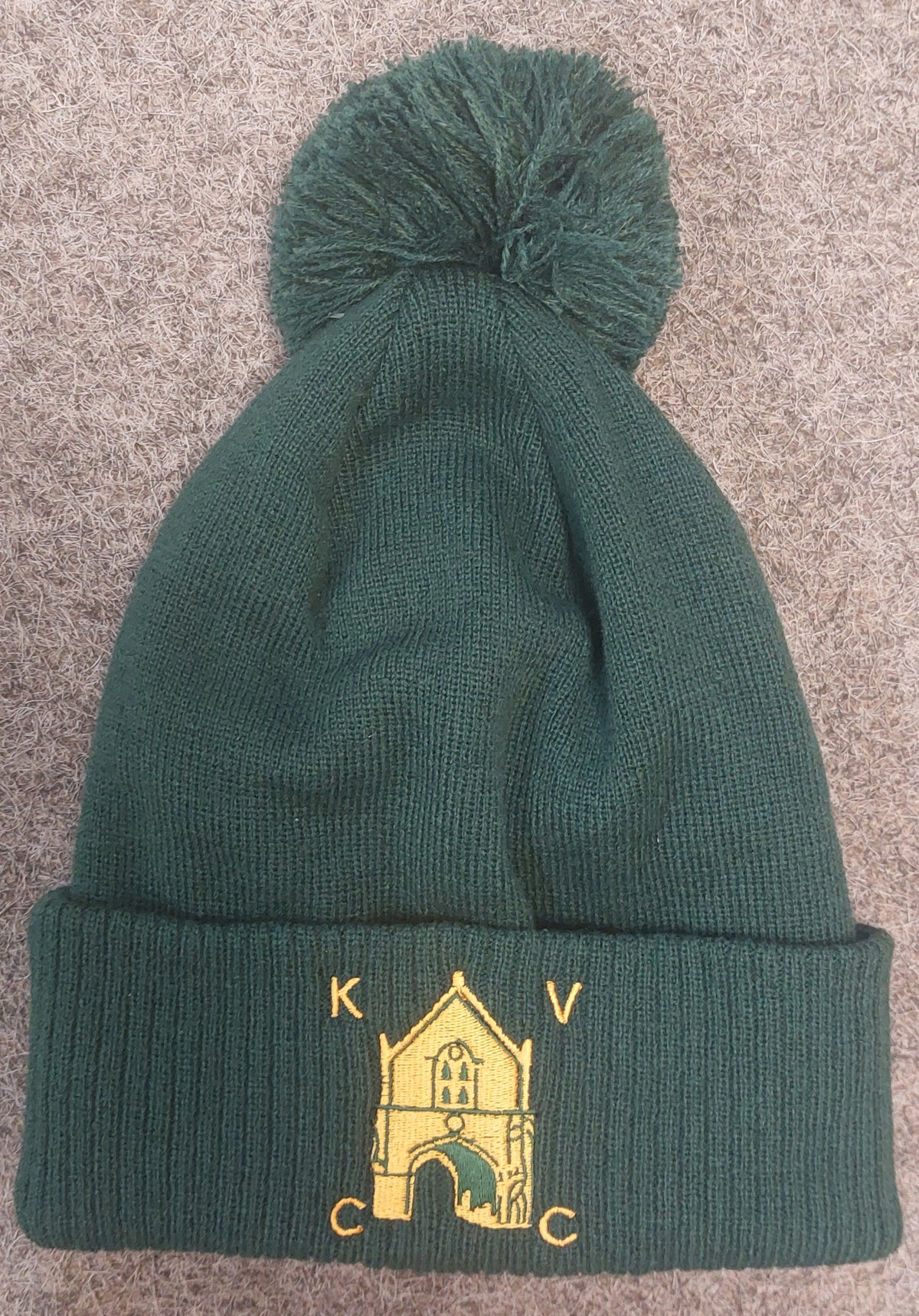 The Kingswood Village CC Thermal Beanie by Martin Berrill Sports is a dark green beanie with a pom-pom and a yellow embroidered club logo with "K V C C" on the cuff, shown on a textured brown surface.