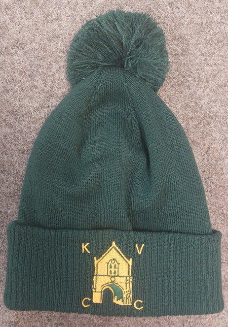 The Kingswood Village CC Thermal Beanie by Martin Berrill Sports is a dark green beanie with a pom-pom and a yellow embroidered club logo with "K V C C" on the cuff, shown on a textured brown surface.